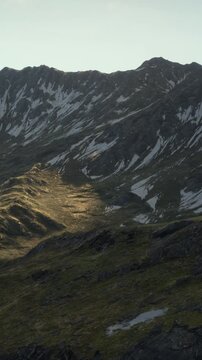 In the wilderness, mountains rise with rocky surfaces and patches of snow. Sunlight touches the land. This scene showcases the rugged beauty of nature during late afternoon light.