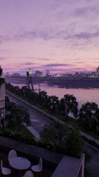 Clouds fill the sky as the sun sets, creating a colorful display. The river reflects the changing light while city buildings are visible in the background. Trees line the riverbank.