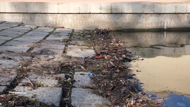 Debris, dry leaves, and litter accumulate along a boat ramp or concrete waterway edge. Concept of urban waterway neglect, pollution, drainage maintenance, and environmental cleanup issues.