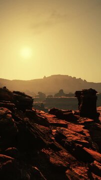 The sun sets behind distant hills, casting a warm glow on rocky terrain. Shadows stretch across the ground as the landscape appears rugged and dry.