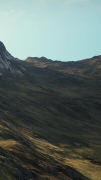 Mountains with rocky surfaces and grassy hills under a clear sky. The sun shines on the landscape highlighting the natural features and colors. It is midday.