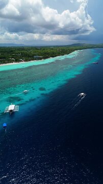 Vertical drone orbits and tracks boats, revealing Dumaluan Beach, coral reef, and blue coastal water in Panglao, Bohol