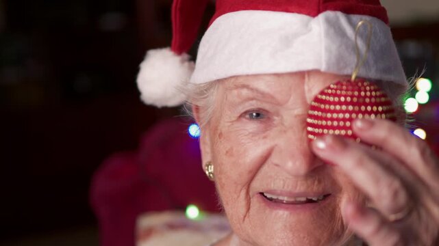 Cheerful senior woman with a santa hat having fun during the christmas holidays, holding a red bauble over her eye and smiling happily at the camera with festive lights in the background