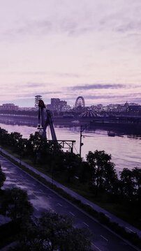 Workers are setting up heavy equipment next to the river. The city skyline is visible in the background along with lights from the amusement park. It is early morning and the sky looks gray.
