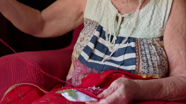 Senior woman wearing a santa hat happily sewing a handmade christmas decoration, carefully crafting a holiday ornament with needle and thread while sitting comfortably in a red armchair at home