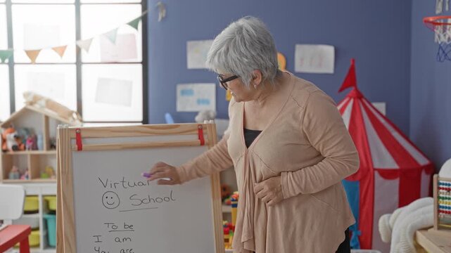 Woman points purple marker at whiteboard in classroom with toys and bunting, teaching a virtual school lesson and writing simple verbs; dedication.