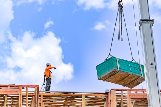 Low angle view of a construction crane lifting a large bundle of green plywood panels to a worker on top of a residential building wood framing structure
