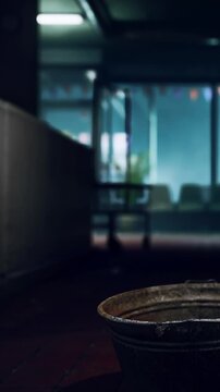 A metal bucket sits on the floor in an empty room. Soft light comes from the windows, creating shadows. Chairs are lined up in the background. The atmosphere is quiet and still.