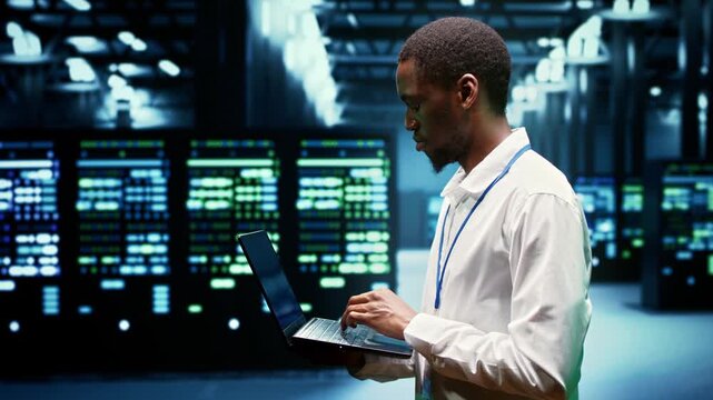 African American engineer walks among electronics in a high tech facility, overseeing computing resources, data storage equipment and network operations during server network.
