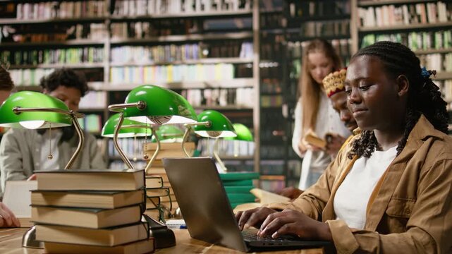 Woman learner studying on laptop and typing beside books, many textbooks on shared wooden table in library, classmates and bookshelves blurred in background, for education technology.