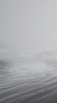 Fog blankets the water as it meets the distant mountains. This scene captures the quiet of early morning with soft reflections on the surface and a gray sky above.