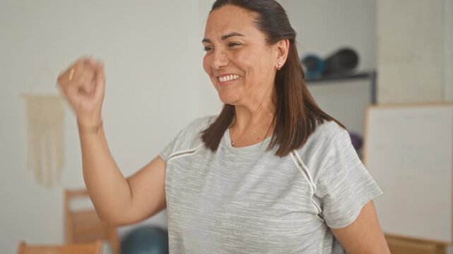 Mature woman raising her arm in a bright yoga studio during gentle training and stretching while smiling, exercise props and whiteboard visible; wellness balance joy.