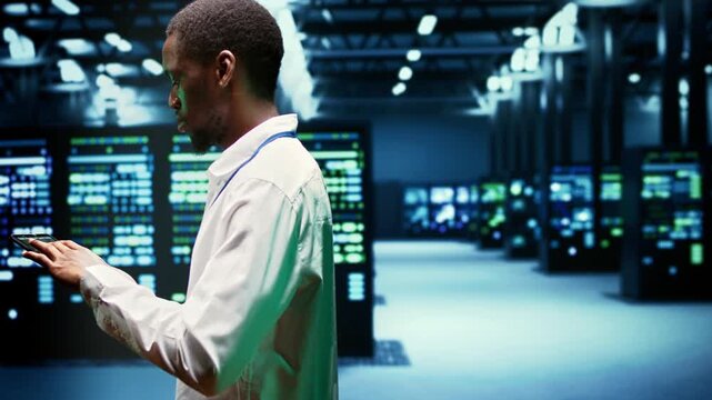 African American IT administrator walks among server clusters in a data center, overseeing computing resources, data storage equipment and network operations during vestigating server clusters.