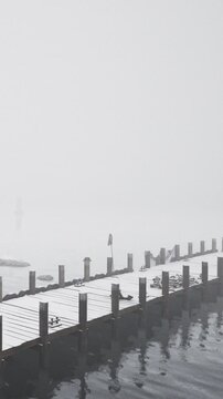 A dock stands in fog with light snow covering its surface. The water reflects the dock and fog. No people are visible. It is early morning and the scene appears quiet and still.