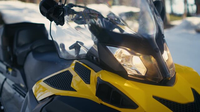 A yellow snowmobile sits in soft spring snow with headlights on and the front windshield in view. Ideal for vehicle, transport, winter and adventure content.