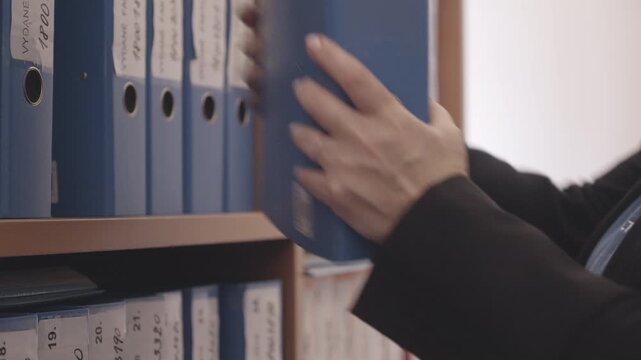 Side profile of an administrative worker replacing an updated binder into office shelving after filing documents. Ideal for records management and business workflow themes.