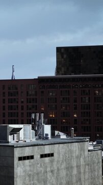 Tall buildings crowd the skyline as dusk arrives. Some windows glow with light while others remain dark. Various rooftop structures are visible in the foreground.