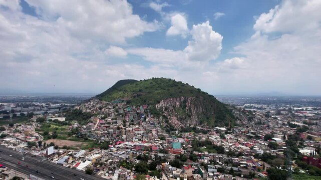 Drone shot approaching Cerro Gordo in Ecatepec, showcasing the mountain, surrounding neighborhoods, highway, urban landscape and dramatic blue sky with white clouds.
