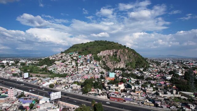 Low altitude drone flight approaching Cerro Gordo in Ecatepec, revealing the mountain, surrounding neighborhoods, highway and expansive blue sky with dramatic clouds over the city.