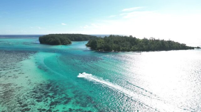 Speedboat carves bright white wake across sunlit turquoise lagoon near Tiahura, Moorea, coral reef patterns visible beneath shallow water surrounding lush green motu. Aerial drone wide shot