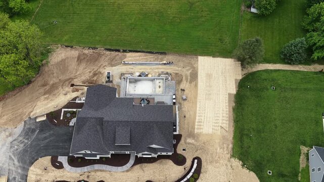 Drone hovers directly above large custom home under construction, new pool basin taking shape beside curved landscaped garden beds and freshly graded green backyard lawn. Aerial drone top down flyover