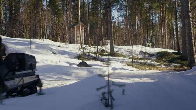 A rider drives a yellow snowmobile past a small forest cabin on a slushy snow track. Ideal for travel, transport, winter adventure and outdoor lifestyle content.