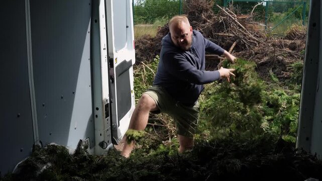Man in work clothes pulls and throws heaps of fresh yard debris from a van interior at an outdoor recycling trip site.