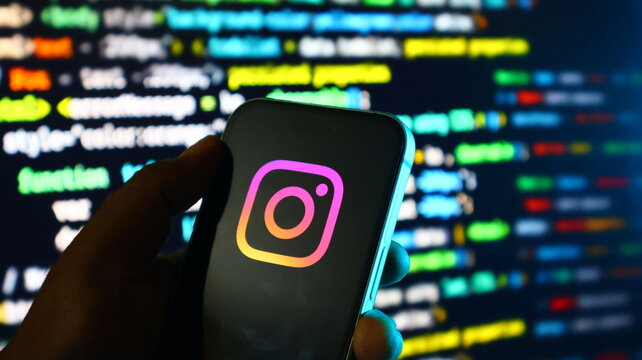 Hand holding smartphone with instagram logo on screen in front of blurred coding background with colorful text