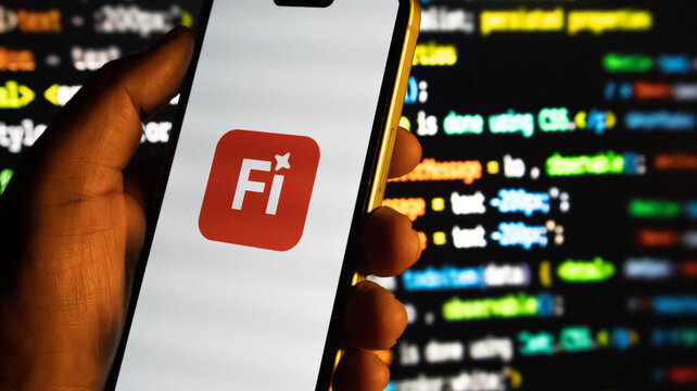 Closeup of a hand holding a smartphone displaying the fi app logo against a background of computer code