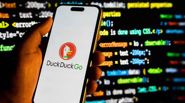 Closeup of a hand holding a smartphone displaying the duckduckgo search engine logo against a background of computer code