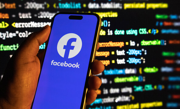 Hand holding a smartphone displaying the facebook logo against a background of computer code