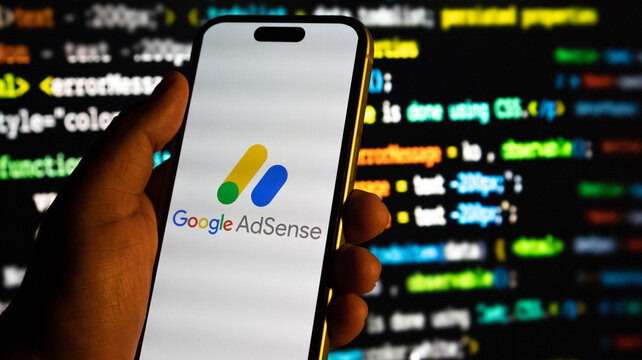 Hand holding a smartphone displaying the google adsense logo against a background of computer code