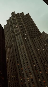 People walk by a tall building in the city at dusk. Some lights are on in the windows. The sky looks gray. The scene shows urban life and tall architecture in the evening.