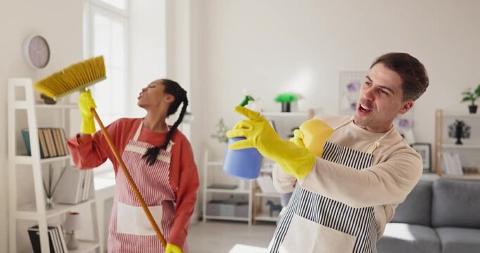 Couple singing and dancing while cleaning home. Man and woman in aprons wear gloves, using broom and spray bottle like mics in a living room. Lighthearted teamwork and cleanliness concept.