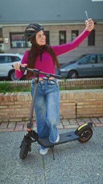 Woman wearing helmet and pink sweater on electric scooter holds smartphone for selfie on street near brick building and parked car; playful joy.