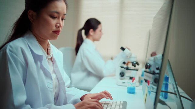 Two female medical scientists in white lab coats collaborating on research. One woman types on a computer while the other examines a sample
