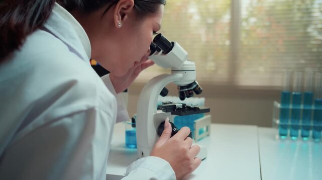 Young Asian woman scientist in a lab coat performing research with a microscope. Professional biochemist analyzing chemical compounds
