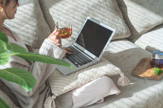A woman sitting on a sofa, sipping tea and looking at her computer.