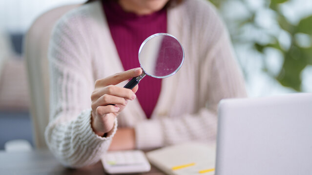 Adult woman holding magnifying glass while studying or researching with laptop and notebook in bright home office