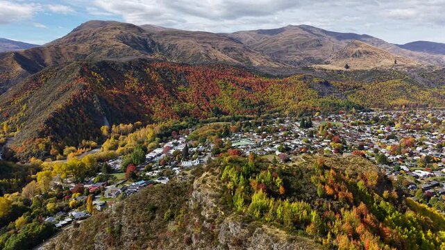Aerial drone video over Arrowtown, New Zealand, with mountains in the foreground, showing the historic South Island town, trees, rooftops, and scenic alpine landscape.