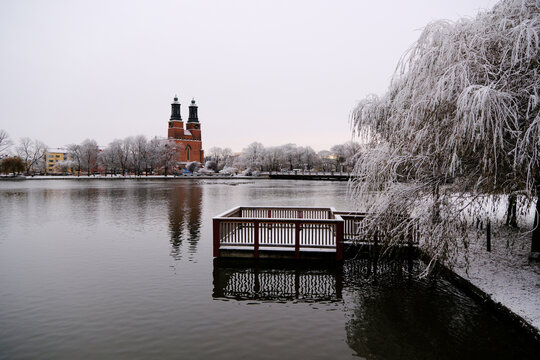 long shot of Eskilstuna River in winter, a snow-covered willow tree and platform in the foreground, with the Klosters Church in the distance reflecting on the calm water.