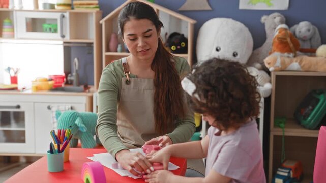 Woman guiding child's drawing with her hand at a table inside a building playroom; nurturing learning joy.