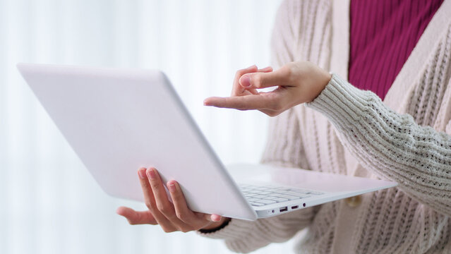 Young Asian woman holding laptop and gesturing while explaining during online education presentation in bright home office