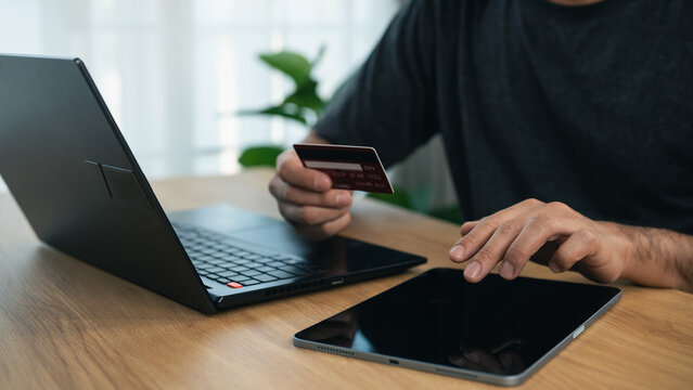 Person making online payment with credit card using laptop and tablet on wooden desk in modern home office setting
