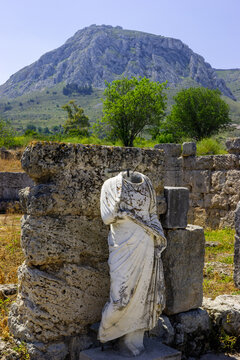 A headless ancient statue stands amidst the ruins of a stone structure, with a rugged, tree-covered mountain rising in the background under a clear blue sky.