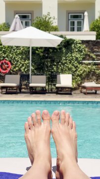 Point of view on female feet enjoying sunbathing by hotel resort swimming pool. Fun during vacation holiday as tourist relaxing on lounge chair by poolside. Barefeet moving. Travel vlogger content