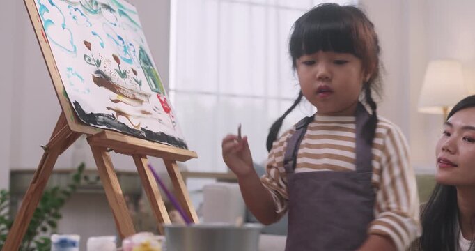 Cute Asian little girl painting on canvas easel with her mother watching at home.