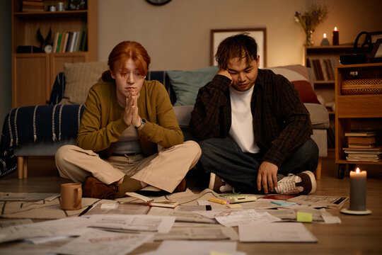 Young adult woman and Asian man sitting on floor, reviewing bills and paperwork, showing financial stress and household budgeting. Useful for debt, inflation, personal finance marketing