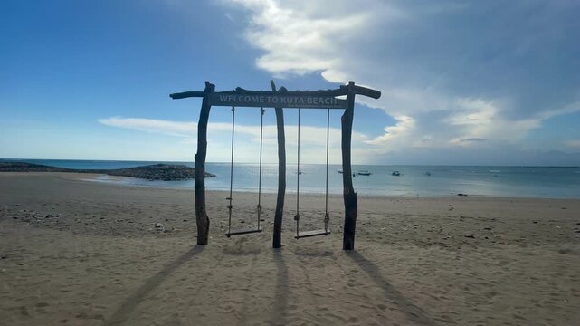 A swing made with woods on the middle at the beach, with word on it say : welcome to kuta beach
