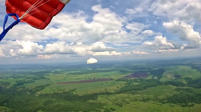 Video footage of a parachute flight over green fields and forests. There are several parachutists in the frame, a cloudy sky and a panoramic landscape.

